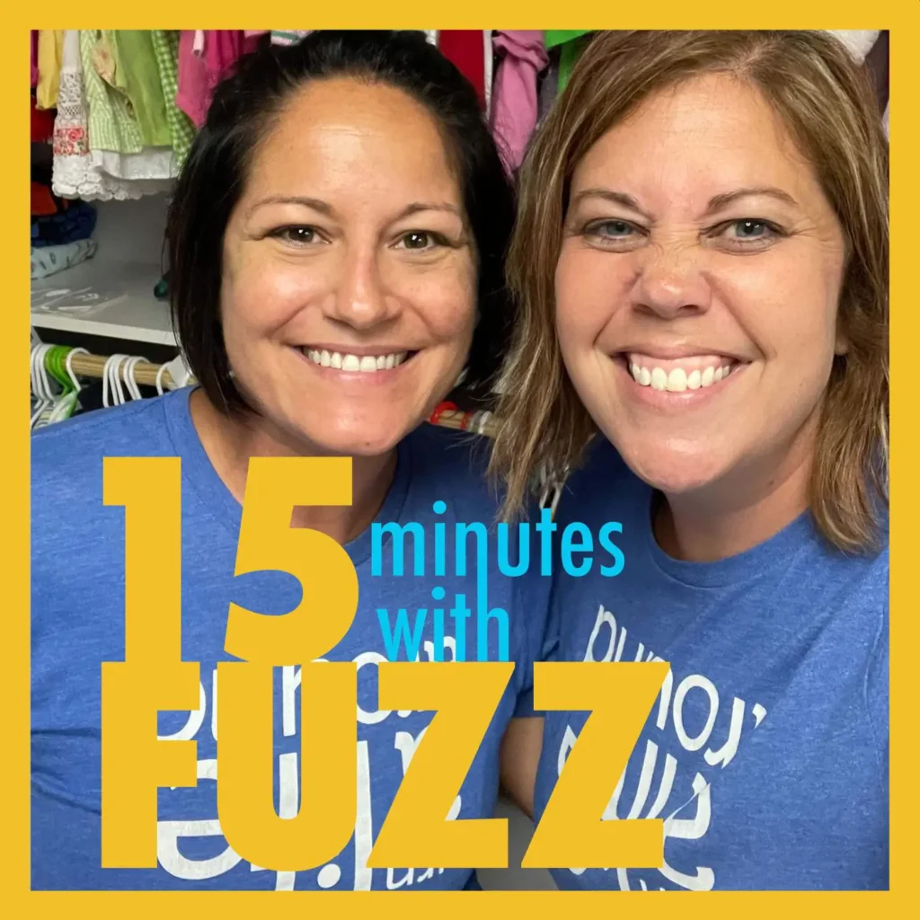 Two women from the Washington County Foster Closet are smiling for the camera. They are both wearing blue t-shirts with white text. The woman on the left has short dark hair, while the woman on the right has shoulder-length light brown hair. Behind them, there are clothes hanging on racks. The image is framed with a yellow border and has the text “15 minutes with FUZZ” in large yellow and blue letters overlaying the bottom portion of the photo.