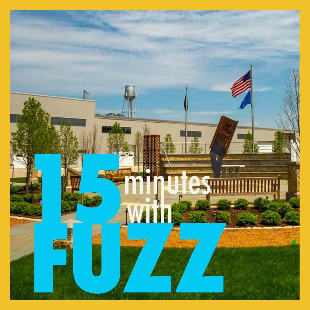 The Wisconsin 9/11 Memorial is pictured on a sunny day with a clear blue sky. The memorial features a landscaped area with bushes, trees, and a stone walkway leading to a large metal sculpture. An American flag and a Wisconsin state flag are flying on flagpoles in the background, along with a large industrial building and a water tower. The image is framed with a yellow border and has the text “15 minutes with FUZZ” in large blue and white letters overlaying the bottom portion of the photo.