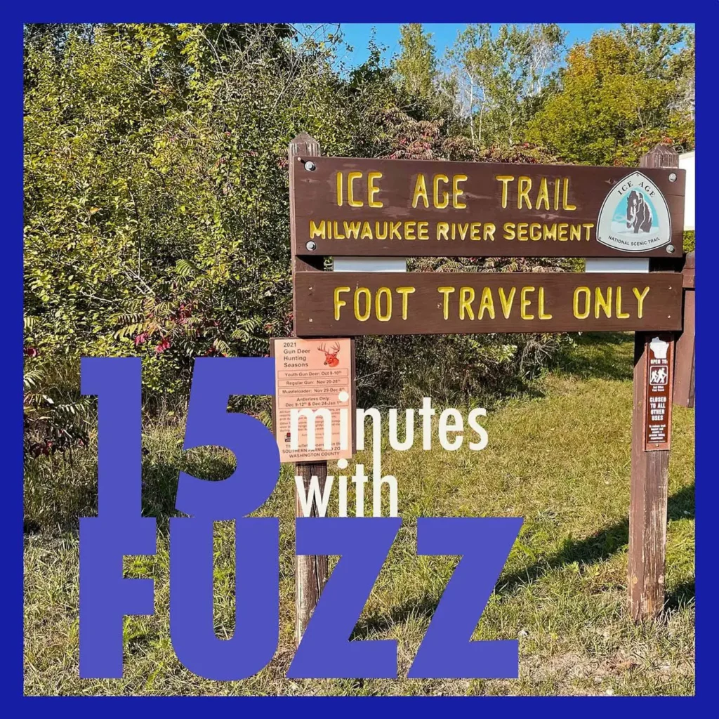 A wooden sign marks the entrance to the Ice Age Trail, Milwaukee River Segment, indicating it is for foot travel only. The sign is situated in a lush, green area with dense trees and bushes in the background. Below the main sign, there is a smaller notice about the deer gun hunting season. The image is framed with a dark blue border and has the text “15 minutes with FUZZ” in large purple and white letters overlaying the bottom portion of the photo.