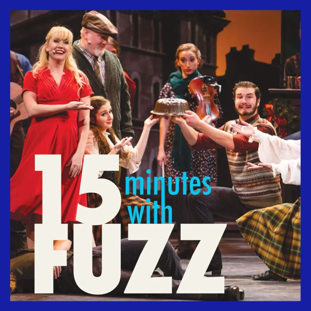 A lively stage performance at the Schauer Arts Center features actors in period costumes. A woman in a bright red dress stands on the left, smiling and gesturing towards a man holding a cake. Other actors, including a man in a flat cap and a woman with a violin, are part of the festive scene, which is set against a backdrop of an old town. The image is framed with a dark blue border and has the text “15 minutes with FUZZ” in large white and blue letters overlaying the bottom portion of the photo.