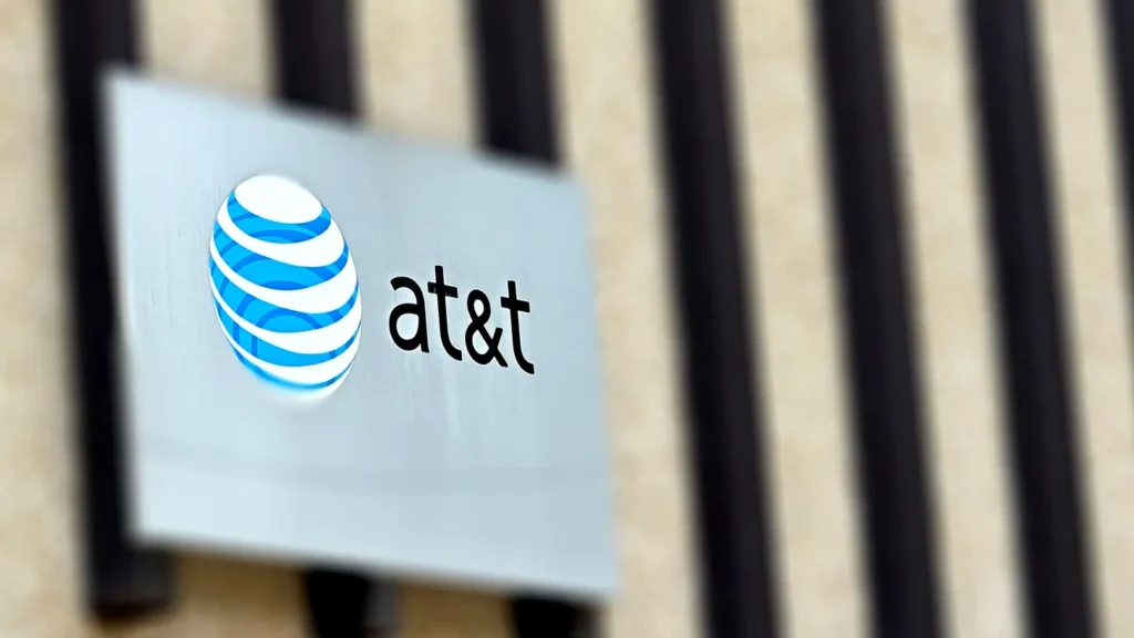 A close-up view of an AT&T sign on the exterior of a building. The sign features the distinctive AT&T logo, a globe with blue and white stripes, positioned on a silver plaque with the company name "at&t" in lowercase letters. The background shows a blurred view of vertical architectural elements, emphasizing the focus on the branded signage. The image captures a modern corporate aesthetic, highlighting the recognizable branding of AT&T.