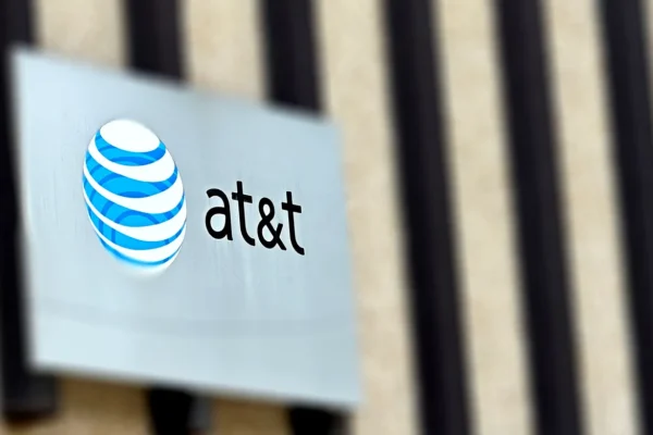 A close-up view of an AT&T sign on the exterior of a building. The sign features the distinctive AT&T logo, a globe with blue and white stripes, positioned on a silver plaque with the company name "at&t" in lowercase letters. The background shows a blurred view of vertical architectural elements, emphasizing the focus on the branded signage. The image captures a modern corporate aesthetic, highlighting the recognizable branding of AT&T.