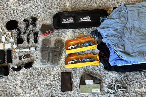 This image features a collection of items laid out on a textured, light-colored carpet, organized for donation. The assortment includes various electronics such as phone chargers, USB cables, and adapters; personal items like sunglasses, a leather wallet, and a pair of running shoes; and several pieces of clothing neatly folded, predominantly t-shirts. Notably, there are also toy cars packaged in yellow boxes, and a weightlifting belt by Schiek. This organized display illustrates a thoughtful decluttering effort, highlighting a range of useful items set to find new homes.