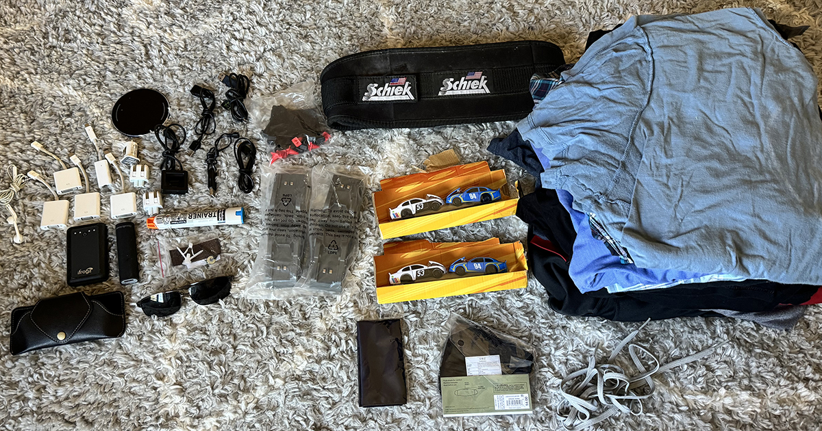 This image features a collection of items laid out on a textured, light-colored carpet, organized for donation. The assortment includes various electronics such as phone chargers, USB cables, and adapters; personal items like sunglasses, a leather wallet, and a pair of running shoes; and several pieces of clothing neatly folded, predominantly t-shirts. Notably, there are also toy cars packaged in yellow boxes, and a weightlifting belt by Schiek. This organized display illustrates a thoughtful decluttering effort, highlighting a range of useful items set to find new homes.