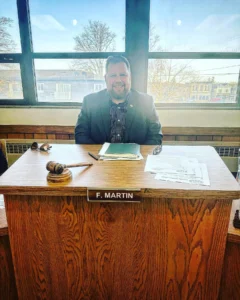 Fuzz Martin sits at the dais in the old Village of Kewaskum village hall. there is a gavel in front of him and his is sitting in front of windows on a sunny day. He is smiling and wearing a sports coat with papers in front of him.