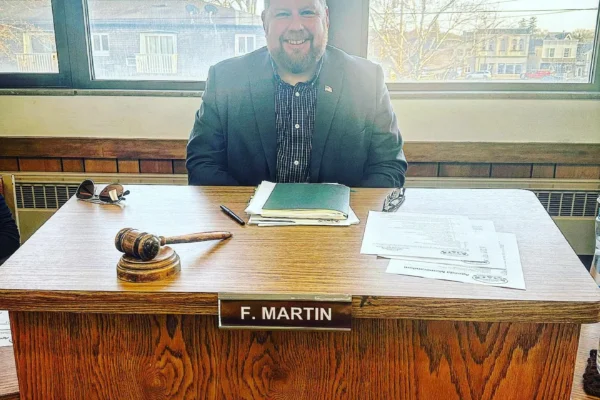 Fuzz Martin sits at the dais in the old Village of Kewaskum village hall. there is a gavel in front of him and his is sitting in front of windows on a sunny day. He is smiling and wearing a sports coat with papers in front of him.
