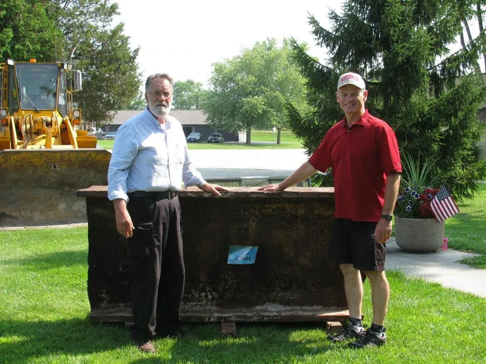 
Two men, Gordon Haberman and Jerry Gosa, stand smiling beside a large piece of steel from the North Tower of the World Trade Center. This relic, serving as a poignant reminder of 9/11, is situated in a park-like setting with lush green grass and a yellow construction vehicle in the background. Gordon, on the left, wears a formal light blue button-up shirt and dark trousers, while Jerry, on the right, is dressed casually in a red polo shirt and dark shorts, donning a Wisconsin baseball cap. A small American flag arrangement adds a touch of patriotic spirit to the scene.