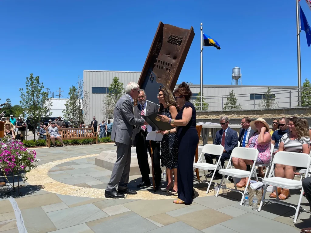 
At the dedication ceremony of the Wisconsin 9/11 Memorial & Education Center, Governor Tony Evers is pictured engaging warmly with Gordon, Kathy, and Julie Haberman. The scene is set under a clear blue sky, with attendees seated in white chairs around a paved area where the ceremony is taking place. Behind the speakers, a significant piece of rusted steel from the World Trade Center stands as a focal point. Spectators, some dressed in suits and others in summer attire, watch attentively in the background, which also features industrial and natural elements, including a water tower. The atmosphere is solemn yet hopeful, marked by the community coming together to honor history and educate future generations.