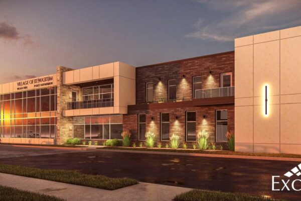 Architectural rendering of the proposed new Village of Kewaskum building. The modern, two-story brick and stone structure features large windows on both floors. Landscaping includes grass and small shrubs. The building is shown at dusk, with interior and exterior lighting illuminating the space. The "Village of Kewaskum" signage is prominently displayed on the front facade.