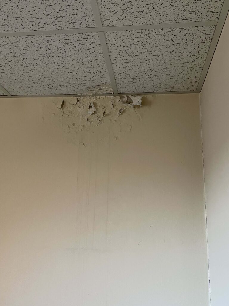 White wall and heavily textured ceiling with visible damage. The ceiling has numerous cracks, and pieces of the texture are flaking off and crumbling. Some debris from the deteriorating ceiling can be seen on the floor below.