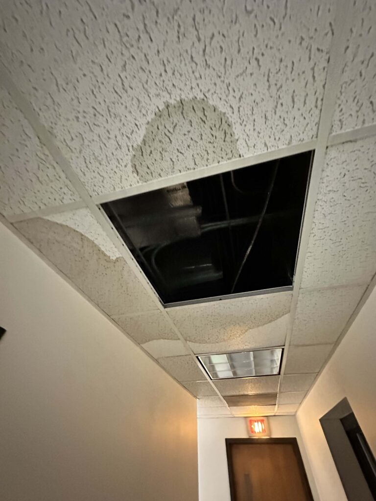 Ceiling of a room with white textured paint or plaster that has numerous cracks and signs of water damage. A dark rectangular opening, likely an access panel, reveals exposed ductwork or plumbing above the damaged ceiling.