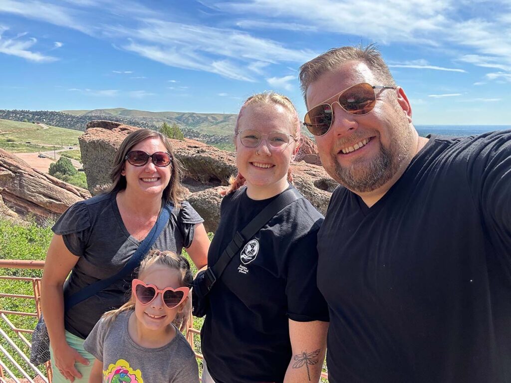 A cheerful family of four is posing for a selfie on a sunny day, set against the backdrop of the rocky, grass-covered hills of Red Rocks Amphitheatre. From left to right: a smiling woman with brown hair wearing sunglasses and a dark gray top, a young woman with blonde hair wearing a black T-shirt, a smiling man with light brown hair wearing sunglasses and a black T-shirt, and a young girl with brown hair wearing heart-shaped sunglasses and a gray T-shirt with a colorful print. They all look happy and relaxed, enjoying the scenic view and pleasant weather.
