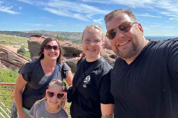 A cheerful family of four is posing for a selfie on a sunny day, set against the backdrop of the rocky, grass-covered hills of Red Rocks Amphitheatre. From left to right: a smiling woman with brown hair wearing sunglasses and a dark gray top, a young woman with blonde hair wearing a black T-shirt, a smiling man with light brown hair wearing sunglasses and a black T-shirt, and a young girl with brown hair wearing heart-shaped sunglasses and a gray T-shirt with a colorful print. They all look happy and relaxed, enjoying the scenic view and pleasant weather.