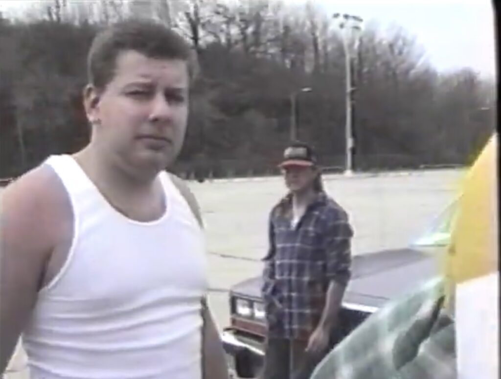 The image depicts a scene from a high school senior skip day in 1996. It features two young men in a parking lot. The man in the foreground is dressed in a white tank top and appears to be talking or interacting with someone off-camera, giving a candid expression. In the background, his friend, wearing a plaid shirt and a baseball cap, leans casually against an older model car. The setting is an outdoor parking area with trees and a clear sky, capturing a relaxed, nostalgic moment from their youth.