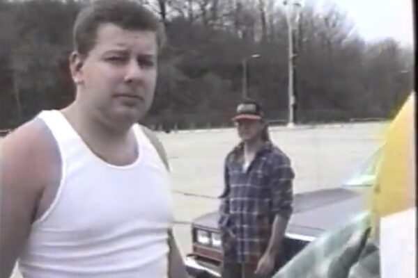 The image depicts a scene from a high school senior skip day in 1996. It features two young men in a parking lot. The man in the foreground is dressed in a white tank top and appears to be talking or interacting with someone off-camera, giving a candid expression. In the background, his friend, wearing a plaid shirt and a baseball cap, leans casually against an older model car. The setting is an outdoor parking area with trees and a clear sky, capturing a relaxed, nostalgic moment from their youth.