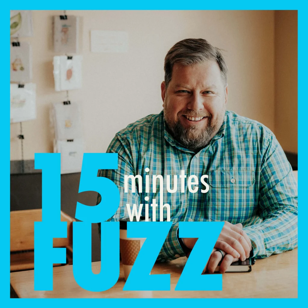 Fuzz Martin is wearing a blue and green plaid shirt and sitting at a table and smiling at the camera. He has a notebook and a coffee cup in front of him. The background features artwork on the wall and a light-colored interior. The image is framed with a light blue border and has the text “15 minutes with FUZZ” in large blue and white letters overlaying the bottom portion of the photo.