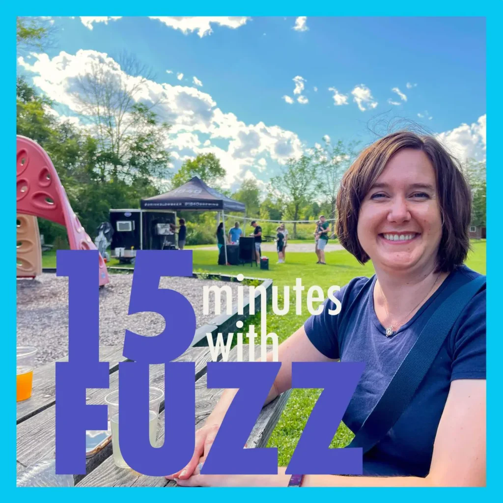 A woman with short brown hair is smiling while sitting at a picnic table at an outdoor event. She is wearing a dark blue t-shirt and has a crossbody bag strap across her shoulder. In the background, there is a playground with a large pink slide, a tent with people gathered around it, and a lush green landscape under a blue sky with scattered clouds. The image is framed with a light blue border and has the text “15 minutes with FUZZ” in large purple and white letters overlaying the bottom portion of the photo.