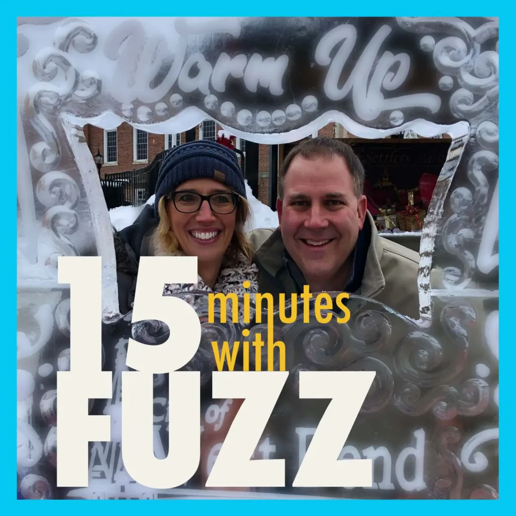 A man and woman are smiling together, framed by an ice sculpture that reads “Warm Up.” The woman is wearing a blue beanie, glasses, and a warm coat, while the man is dressed in a beige jacket. They are standing outside in a snowy environment with a brick building in the background. The image is framed with a light blue border and has the text “15 minutes with FUZZ” in large white and yellow letters overlaying the bottom portion of the photo.