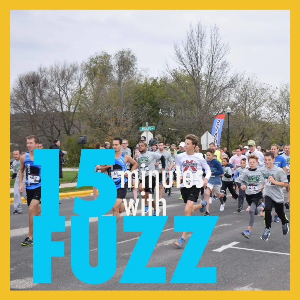 A group of runners are starting a race at the KEYS Scholarship Run in Kewaskum. The participants, dressed in various athletic outfits and numbered bibs, are running on a road with trees and a street sign in the background. An orange flag marked “START” is visible in the crowd. The sky is overcast. The image is framed with a yellow border and has the text “15 minutes with FUZZ” in large blue and white letters overlaying the bottom portion of the photo.