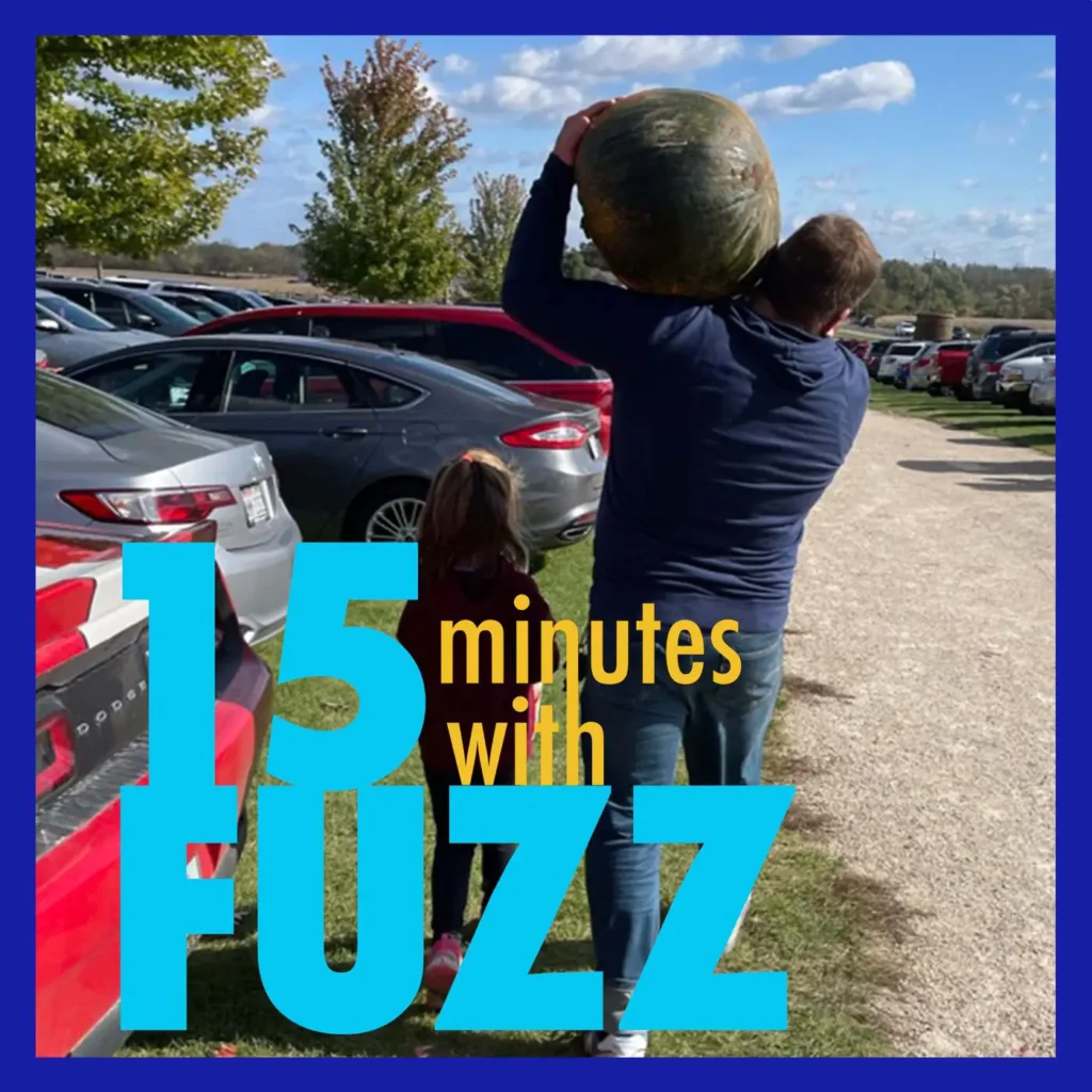 A man is carrying a large pumpkin on his shoulder while walking through a parking lot filled with cars. A young girl, holding his hand, walks beside him. They are at an outdoor event on a sunny day with a clear blue sky and trees in the background. The image is framed with a dark blue border and has the text “15 minutes with FUZZ” in large blue and yellow letters overlaying the bottom portion of the photo.
