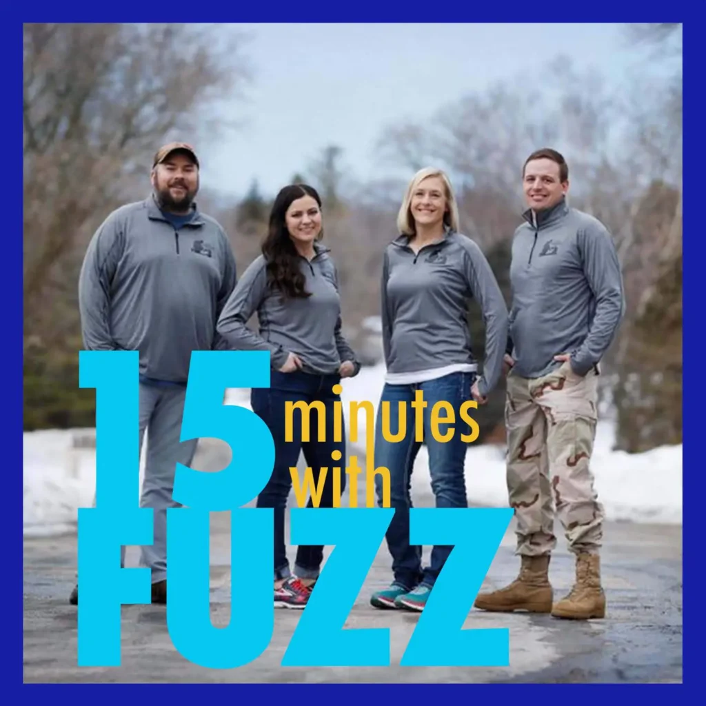 Four people are standing together outside on a snowy day, wearing matching gray jackets with logos on them. From left to right, the group includes a bearded man in a cap, a woman with long dark hair, a blonde woman, and a man in camouflage pants and brown boots. They are all smiling and have their hands in their pockets. The background features a winter landscape with bare trees. The image is framed with a dark blue border and has the text “15 minutes with FUZZ” in large blue and yellow letters overlaying the bottom portion of the photo.