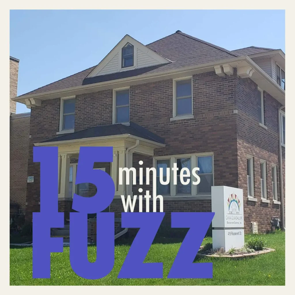 A two-story brick building with a gabled roof is pictured on a sunny day. The building has multiple windows and a small covered porch at the entrance. A sign in front of the building reads “Casa Guadalupe Education Center, Inc.” with a colorful logo. The grass around the building is neatly trimmed. The image is framed with a white border and has the text “15 minutes with FUZZ” in large purple and white letters overlaying the bottom portion of the photo.