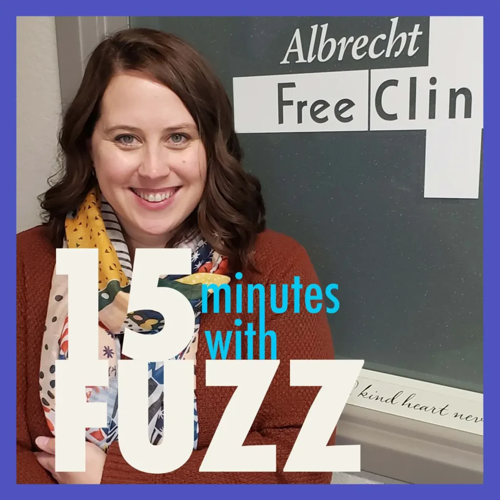 A woman with shoulder-length brown hair is smiling and standing in front of a door with a sign that reads “Albrecht Free Clinic.” She is wearing a colorful scarf and a brown sweater. The image is framed with a dark blue border and has the text “15 minutes with FUZZ” in large white and blue letters overlaying the bottom portion of the photo.