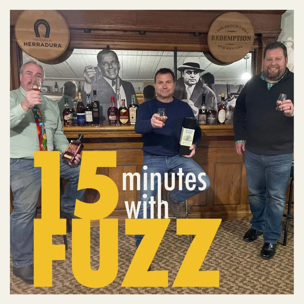 Three men are standing at a bar, each holding a glass of liquor. The man on the left is wearing a green shirt and a colorful tie, the man in the center is in a dark blue sweater and jeans holding a bottle of whiskey, and the man on the right is wearing a dark sweater and jeans. Behind them, the bar is stocked with various bottles, and there are black-and-white photos of famous historical figures on the wall. The setting appears cozy with wooden paneling. The image is framed with a white border and has the text “15 minutes with FUZZ” in large yellow and white letters overlaying the bottom portion of the photo.