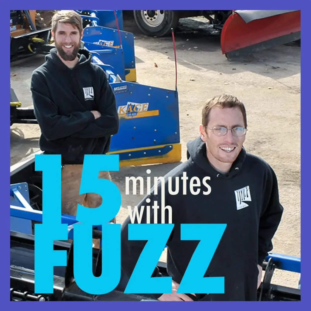 Two men wearing black hoodies with a company logo are standing outdoors near blue and yellow snow removal equipment. The man on the left has a beard and his arms crossed, while the man on the right is smiling with his hands in his pockets. They are standing on a concrete surface with more equipment and a truck visible in the background. The image is framed with a purple border and has the text “15 minutes with FUZZ” in large blue and white letters overlaying the bottom portion of the photo.