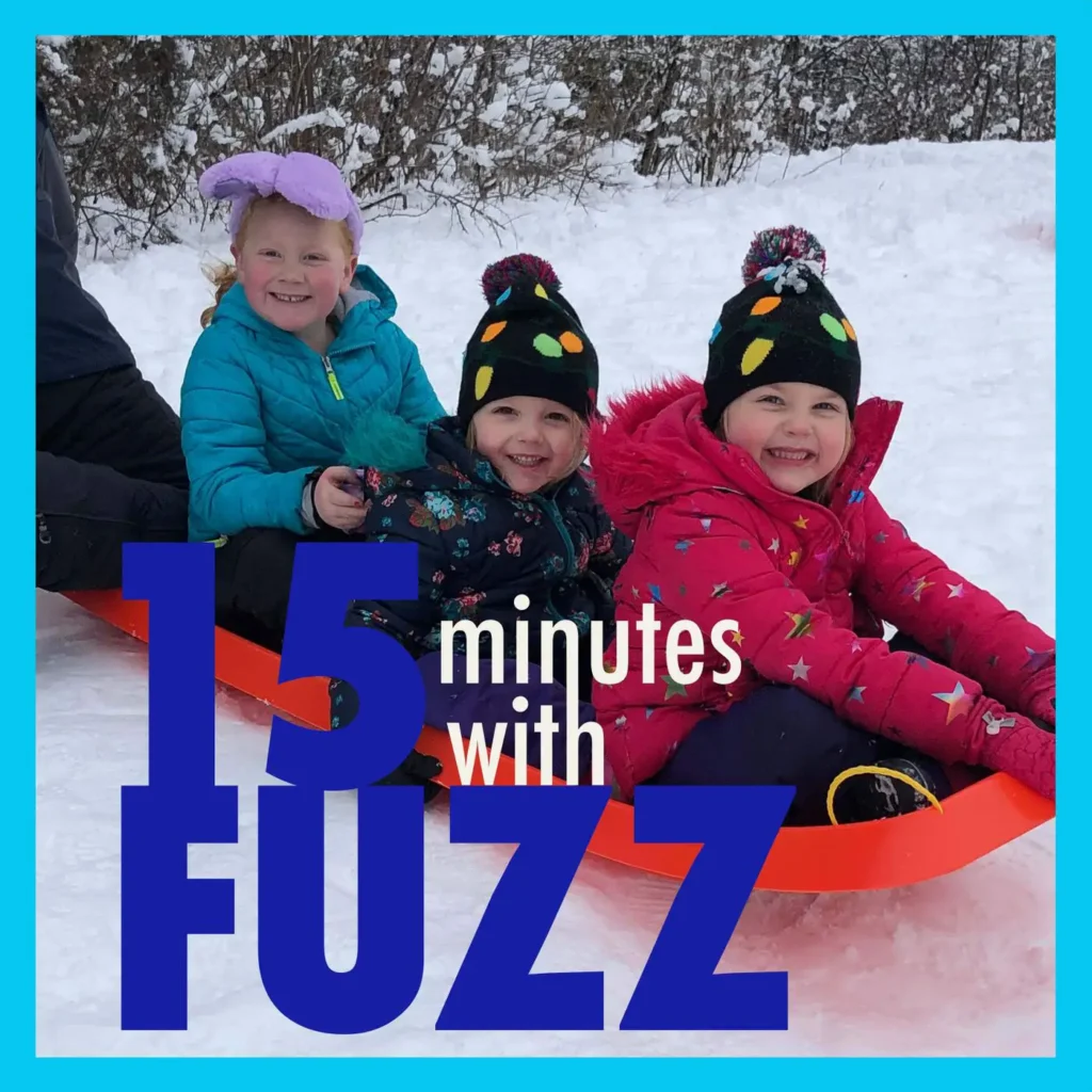 Three young children are sitting together on an orange sled in the snow, smiling at the camera. They are dressed in winter coats and hats, with the child on the left wearing a turquoise jacket and purple bow hat, the middle child wearing a floral jacket and black hat with colorful dots, and the child on the right wearing a bright pink jacket and a similar hat. Snow-covered bushes are visible in the background. The image is framed with a light blue border and has the text “15 minutes with FUZZ” in large dark blue and white letters overlaying the bottom portion of the photo.