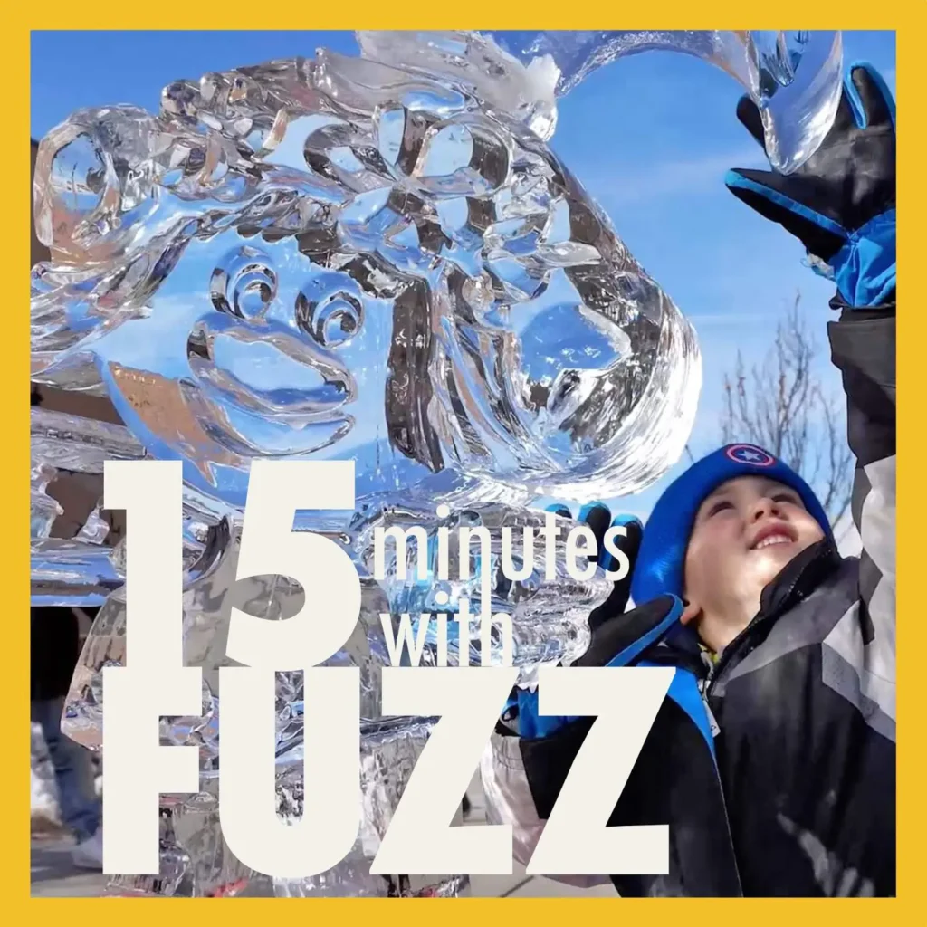 A young child dressed in winter clothing, including a blue hat and gloves, smiles while standing next to a large ice sculpture. The sculpture features intricate designs and a cartoonish face. The child is looking up at the sculpture with excitement. The sky is clear and blue, indicating a bright winter day. The image is framed with a yellow border and has the text “15 minutes with FUZZ” in large white and blue letters overlaying the bottom portion of the photo.