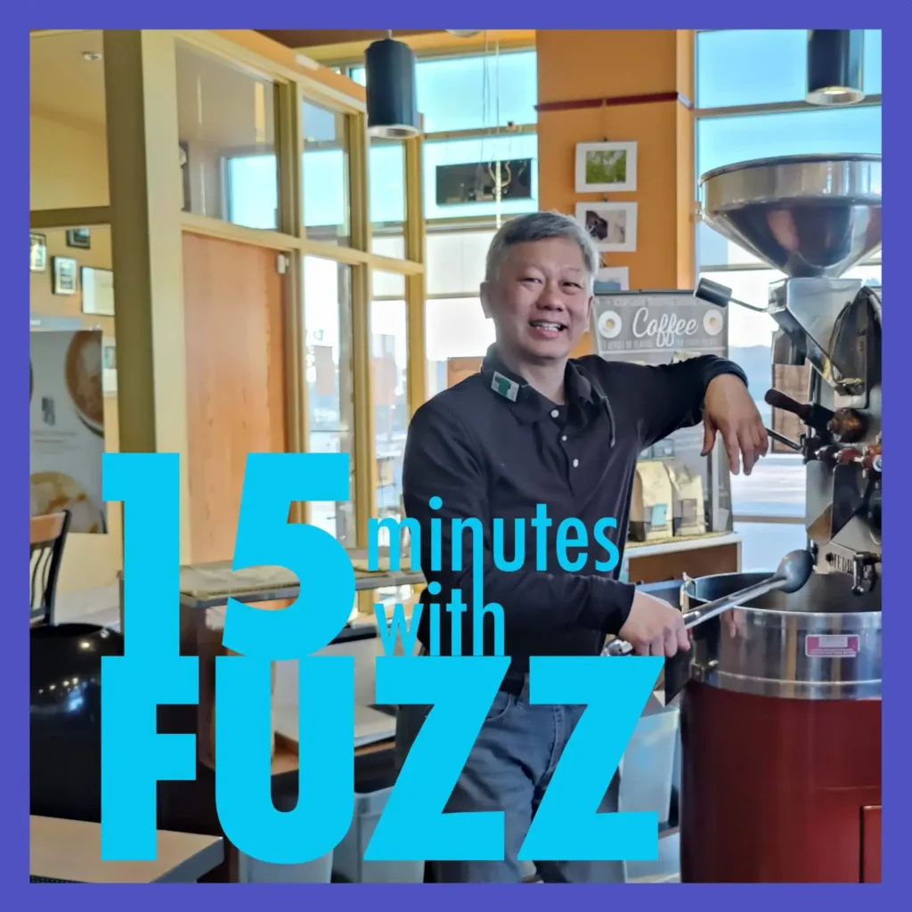 Al Jung is smiling and standing next to a coffee roasting machine inside Dunn Bros Coffee in West Bend. He is wearing a black shirt with a green logo and jeans. The interior of the coffee shop features large windows, wooden furniture, and coffee-related decorations. The image is framed with a purple border and has the text “15 minutes with FUZZ” in large blue and white letters overlaying the bottom portion of the photo.
