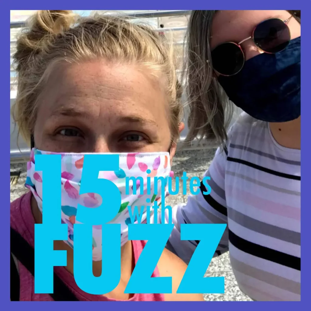 Two women wearing face masks are taking a selfie outdoors. The woman on the left has blonde hair tied up and is wearing a colorful mask with pink and blue patterns, while the woman on the right is wearing sunglasses and a striped shirt with a dark blue mask. They are standing in front of a light-colored background, likely a building or vehicle. The image is framed with a purple border and has the text “15 minutes with FUZZ” in large blue and white letters overlaying the bottom portion of the photo.