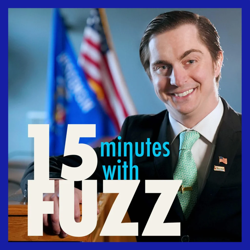 A man in a suit is smiling while standing next to a podium. He is wearing a green patterned tie and has an American flag pin on his lapel. Behind him, the American flag and Wisconsin state flag are visible, slightly out of focus. The man has neatly combed hair and is dressed formally. The image is framed with a dark blue border and has the text “15 minutes with FUZZ” in large white and blue letters overlaying the bottom portion of the photo.