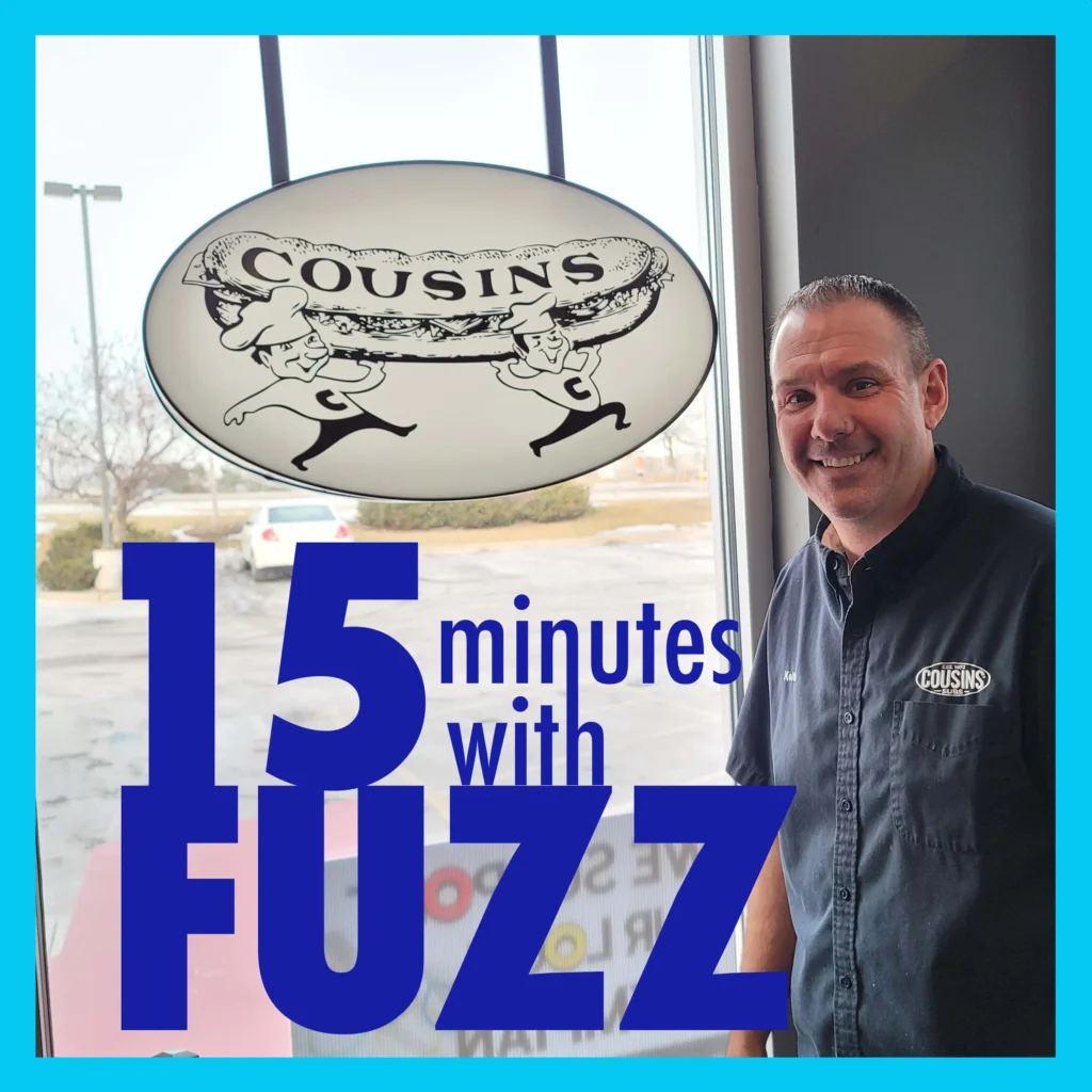 A man is smiling while standing inside a Cousins Subs restaurant. He is wearing a dark shirt with the Cousins Subs logo on it. Behind him, there is a large window with a view of the parking lot and a Cousins Subs sign hanging above. The sign features an illustration of two chefs carrying a large sandwich. The image is framed with a light blue border and has the text “15 minutes with FUZZ” in large dark blue and white letters overlaying the bottom portion of the photo.