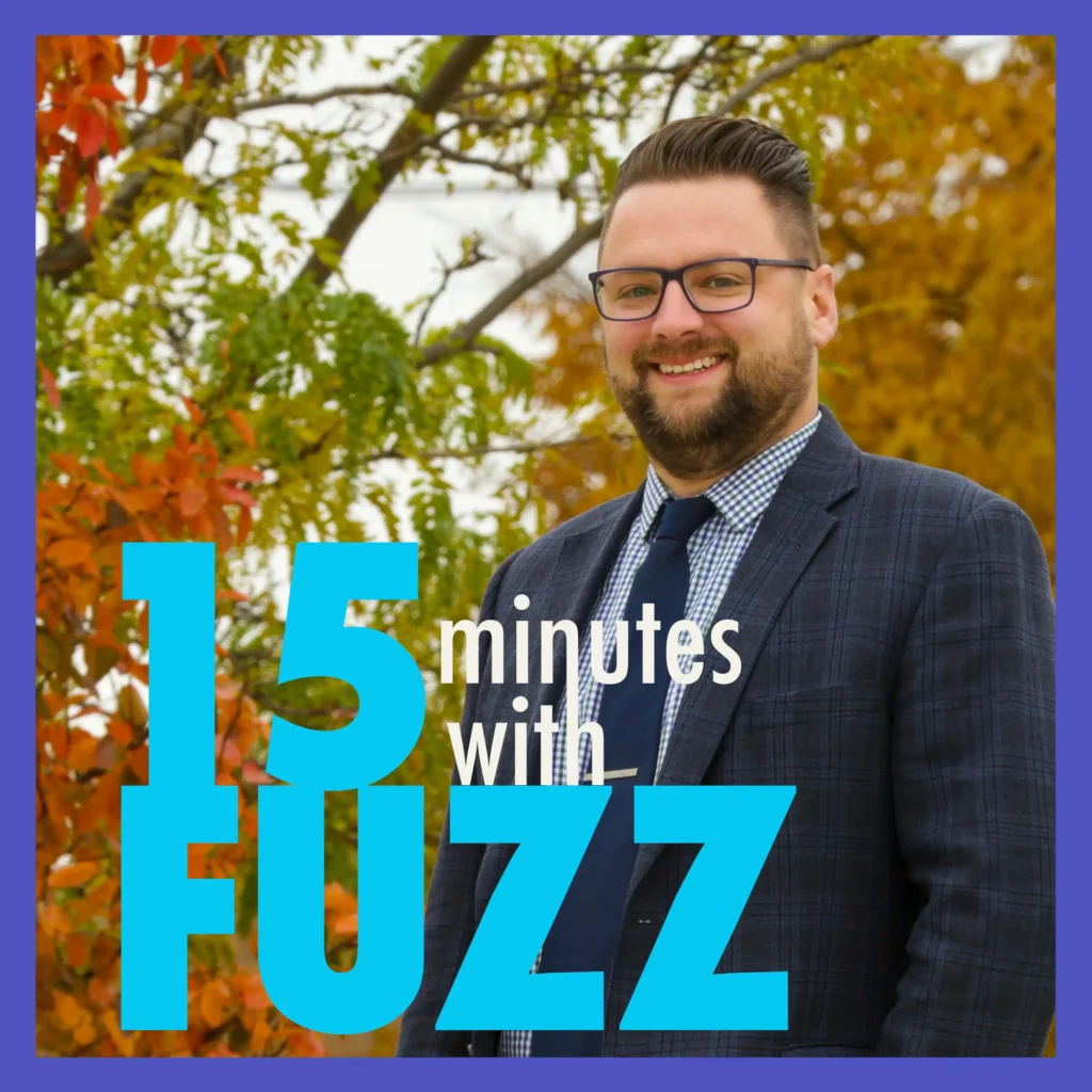 A man with a beard and glasses is smiling while standing outdoors in front of autumn foliage. He is wearing a dark blue plaid blazer, a checkered shirt, and a dark blue tie. The trees behind him display vibrant green, orange, and yellow leaves. The image is framed with a purple border and has the text “15 minutes with FUZZ” in large blue and white letters overlaying the bottom portion of the photo.