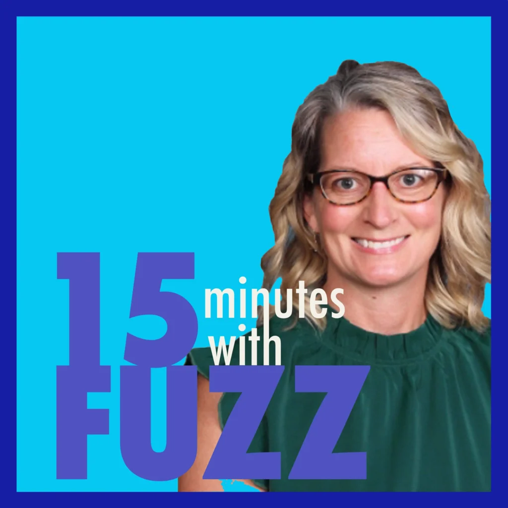 A woman with blonde wavy hair and glasses is smiling at the camera. She is wearing a dark green sleeveless blouse. The background is a solid light blue color. The image is framed with a dark blue border and has the text “15 minutes with FUZZ” in large purple and white letters overlaying the bottom portion of the photo.