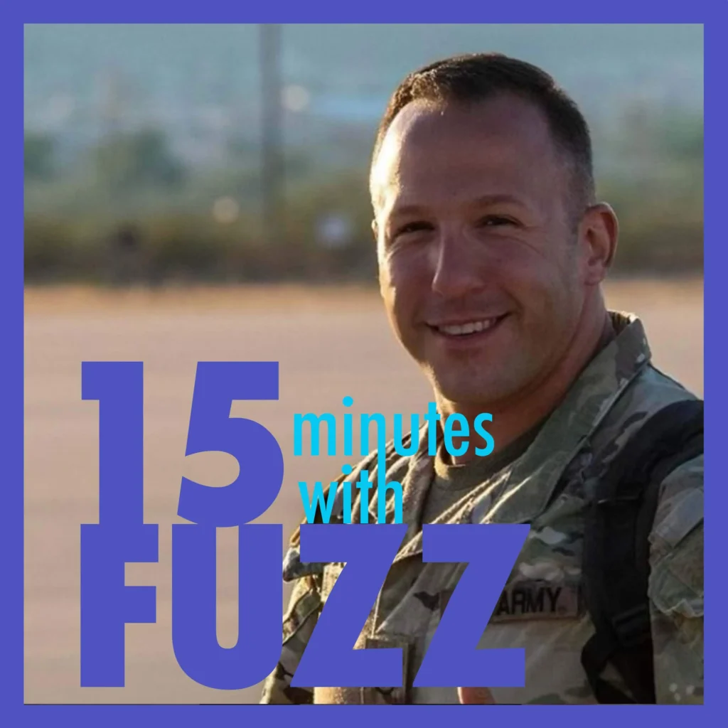 A man in military uniform is smiling at the camera. He is wearing camouflage gear with “Army” written on it and has a backpack on his shoulders. The background is an outdoor setting with blurred landscape details. The image is framed with a purple border and has the text “15 minutes with FUZZ” in large blue and white letters overlaying the bottom portion of the photo.