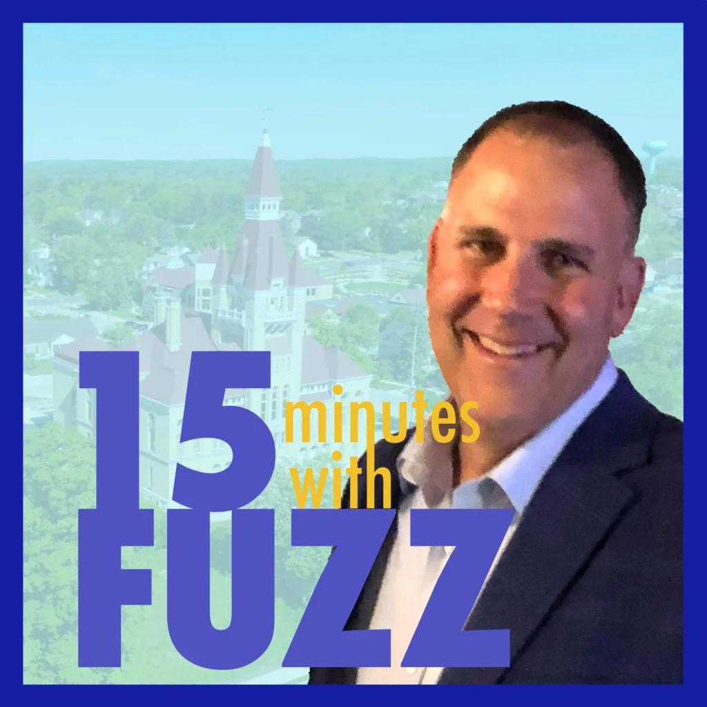 Jay Shambeau is in a suit smiling at the camera with a blurred aerial view of a cityscape in the background, featuring a prominent red-roofed building. He is wearing a blue blazer and a light blue shirt. The image is framed with a dark blue border and has the text “15 minutes with FUZZ” in large purple and yellow letters overlaying the bottom portion of the photo.