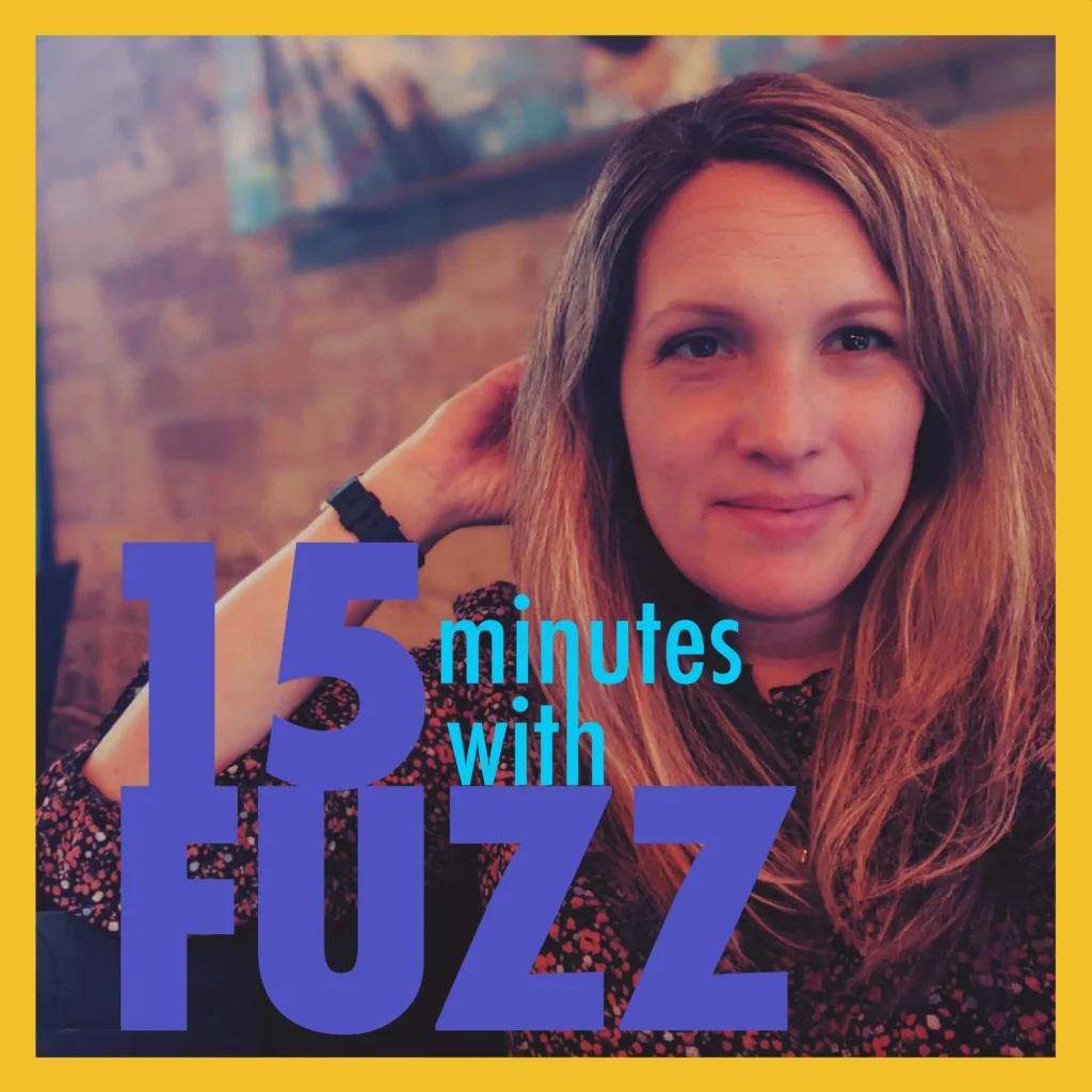 A woman with long, light brown hair is smiling and resting her hand on the back of her head. She is wearing a floral-patterned blouse and sitting indoors, with a blurred background featuring warm tones and artwork on the wall. The image is framed with a yellow border and has the text “15 minutes with FUZZ” in large purple and blue letters overlaying the bottom portion of the photo.