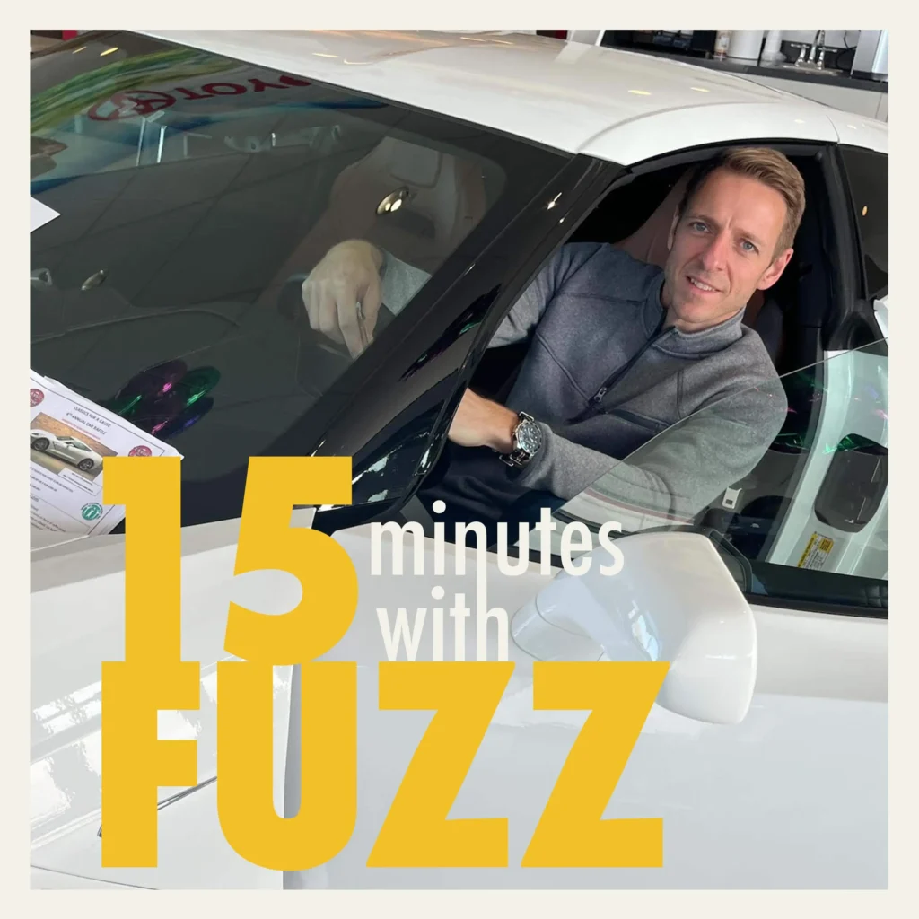 A man is sitting in the driver’s seat of a white car, smiling at the camera. He is wearing a gray zip-up sweater. The car is indoors, likely in a showroom, with a Toyota logo visible in the background. The image is framed with a white border and has the text “15 minutes with FUZZ” in large yellow and white letters overlaying the bottom portion of the photo.
