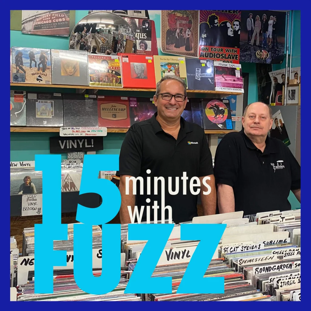 Two men are standing inside a record store, smiling at the camera. The man on the left is wearing a black shirt with a Microsoft logo, and the man on the right is wearing a black shirt with the store’s logo, “The Beat Goes On Records and More.” Behind them, the walls are filled with various vinyl records, posters, and music memorabilia. The image is framed with a dark blue border and has the text “15 minutes with FUZZ” in large blue and white letters overlaying the bottom portion of the photo.
