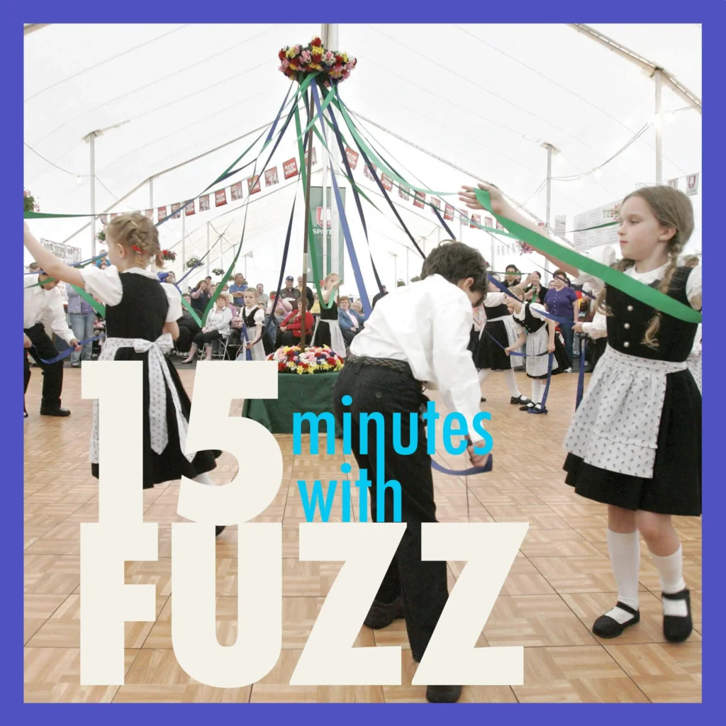The image shows children participating in a traditional Maypole dance at a community event. The children are dressed in traditional German attire, including aprons and dresses for the girls and white shirts with dark pants for the boys. They are holding and weaving colorful ribbons around a Maypole, which is decorated with flowers at the top. The event appears to be taking place inside a large tent with a wooden floor and banners hanging from the ceiling. In the foreground, the text “15 minutes with Fuzz” is prominently displayed in a bold font.