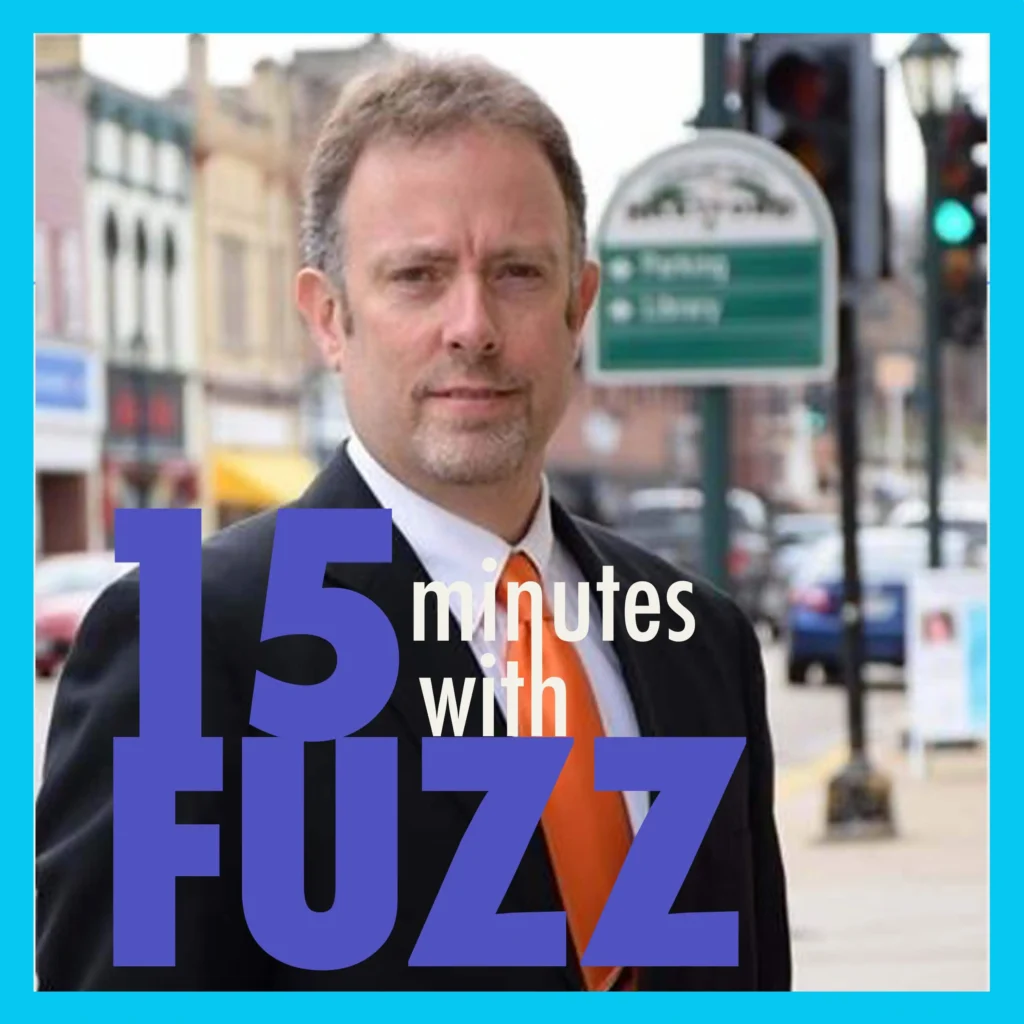 The image features a man in a suit and tie standing on a sidewalk in a small-town setting. The man, dressed in a black suit with an orange tie, is looking directly at the camera with a slight smile. Behind him, there are buildings with storefronts, a street sign, and a few parked cars, indicating a downtown area. The background is slightly blurred, focusing attention on the man. In the foreground, the text “15 minutes with Fuzz” is prominently displayed in a bold font.