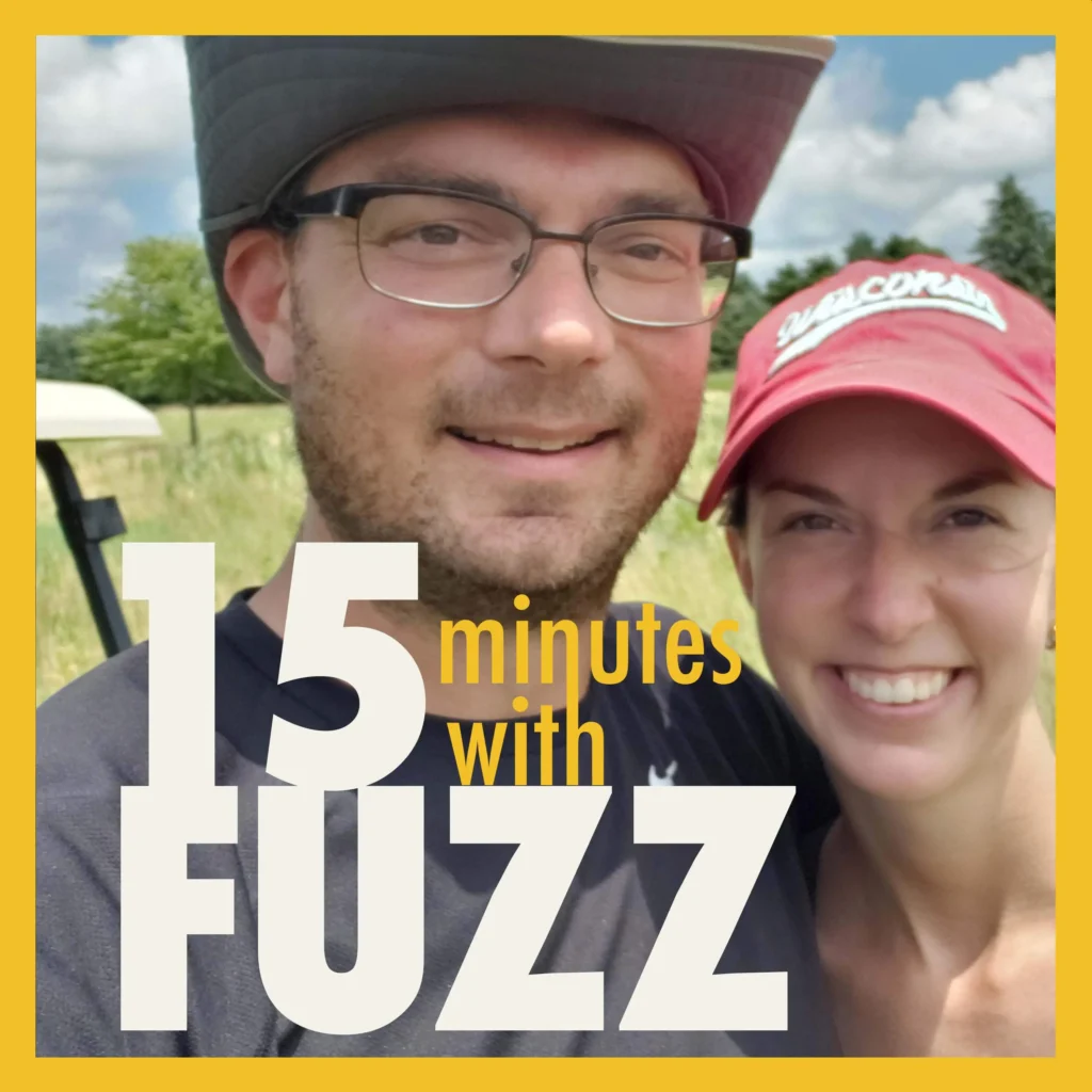 The image shows a smiling man and woman taking a selfie outdoors on a sunny day. The man is wearing a wide-brimmed hat and glasses, while the woman is wearing a red baseball cap with the word “Wisconsin” on it. They are both casually dressed and appear to be enjoying a day out in nature, with green fields and trees in the background. The sky is partly cloudy, adding to the bright and cheerful atmosphere. In the foreground, the text “15 minutes with Fuzz” is prominently displayed in a bold font.