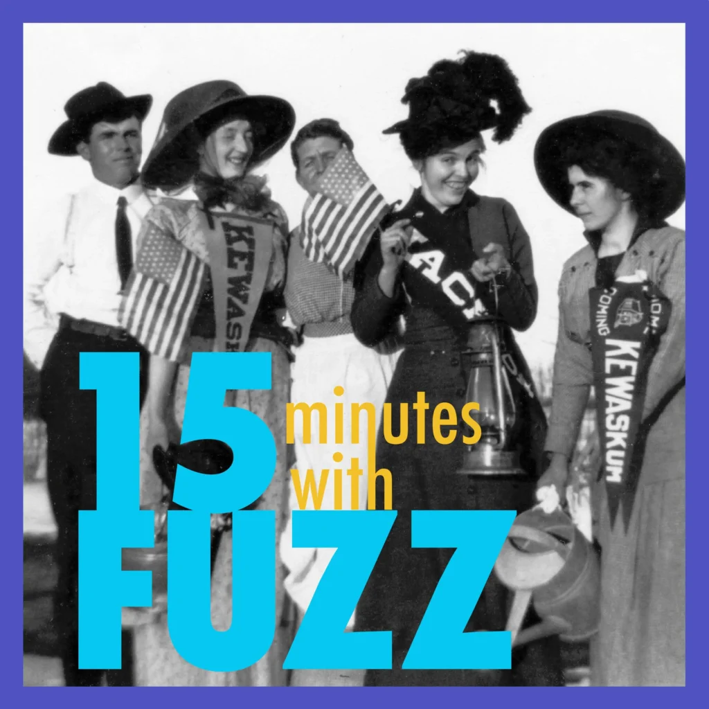 The image is a historical black-and-white photograph featuring a group of men and women dressed in early 20th-century attire. The individuals are wearing wide-brimmed hats, sashes, and holding American flags, giving the scene a patriotic and celebratory atmosphere. Some of the sashes read “Kewaskum,” indicating the location or event they are celebrating. One woman is holding a lantern, while another holds a watering can. The group is smiling and appears to be enjoying the occasion. In the foreground, the text “15 minutes with Fuzz” is prominently displayed in a bold font.