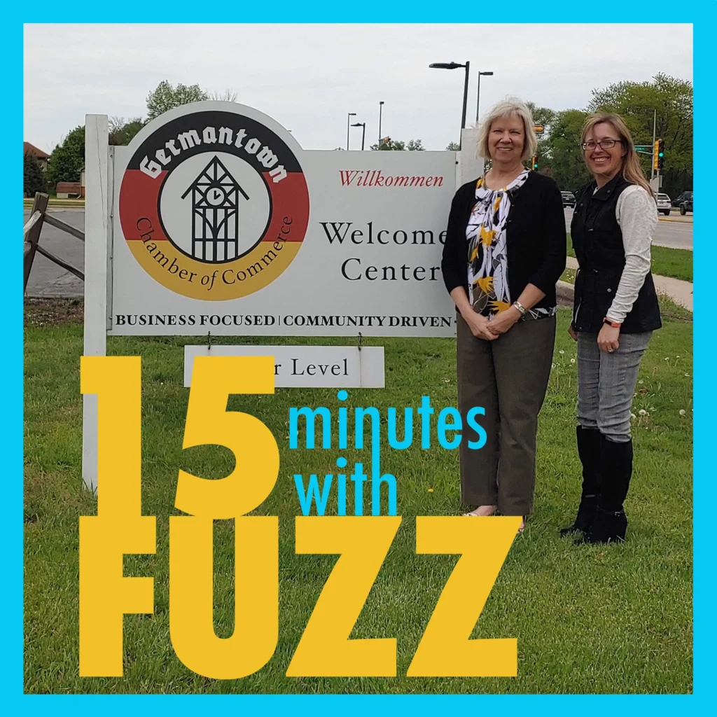 The image features two women standing in front of a sign for the Germantown Chamber of Commerce. The sign reads “Germantown Chamber of Commerce Welcome Center” and includes the phrases “Business Focused” and “Community Driven.” The women are smiling and dressed in business casual attire, with one wearing a black cardigan and the other a black vest over a light-colored top. They are standing on a grassy area near a sidewalk, with trees and a street visible in the background. In the foreground, the text “15 minutes with Fuzz” is prominently displayed in a bold font.
