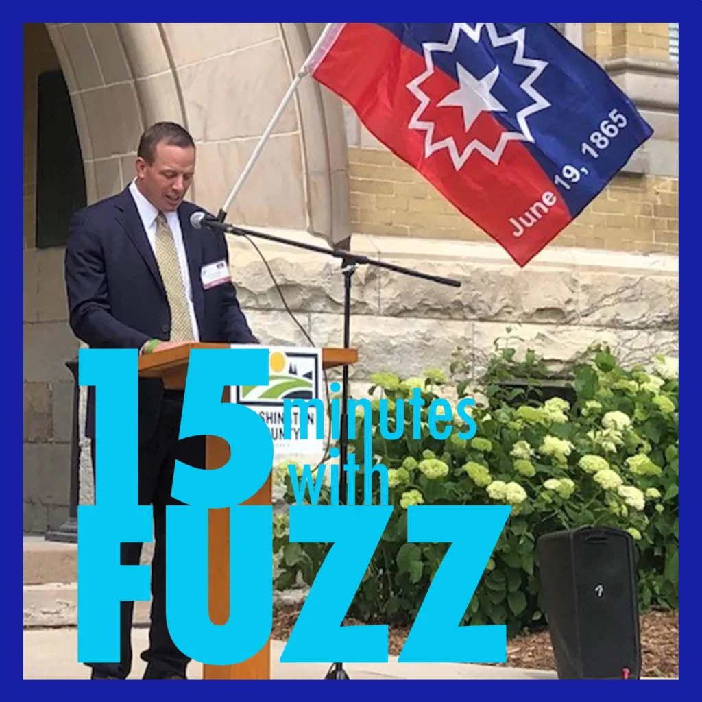County Executive Josh Schoemann is in a suit and tie standing at a podium and speaking into a microphone. He is giving a speech at an outdoor event, with a flag in the background that reads “June 19, 1865,” indicating the celebration of Juneteenth. The flag features a prominent star and colors of red, white, and blue. The podium has a logo for Washington County. Surrounding the area are green plants and flowers, and the backdrop includes a stone building with arched architecture. In the foreground, the text “15 minutes with Fuzz” is prominently displayed in a bold font.