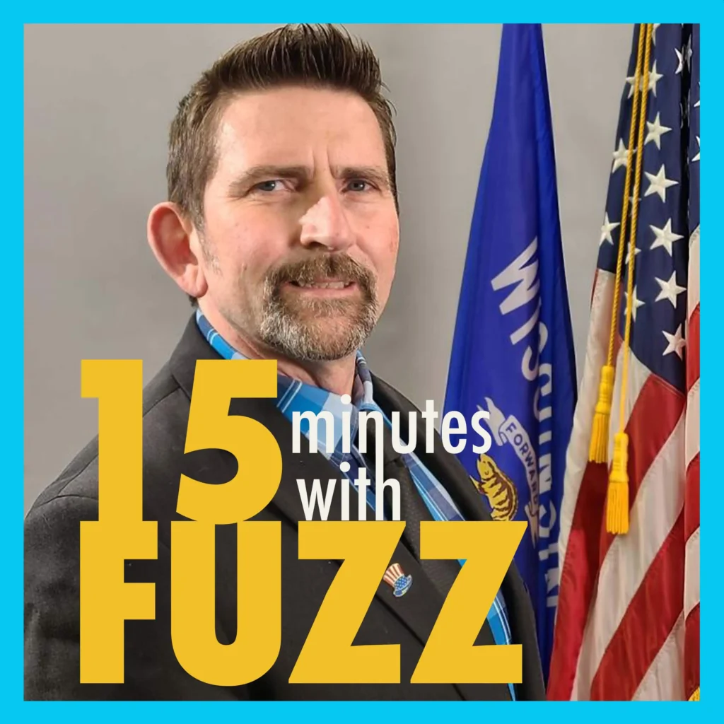 A man with short brown hair and a mustache, dressed in a suit with a blue shirt, stands in front of the Wisconsin and American flags. He is smiling slightly and looking directly at the camera. The background is neutral, drawing focus to the man and the flags. In the foreground, the text “15 minutes with Fuzz” is prominently displayed in a bold font.
