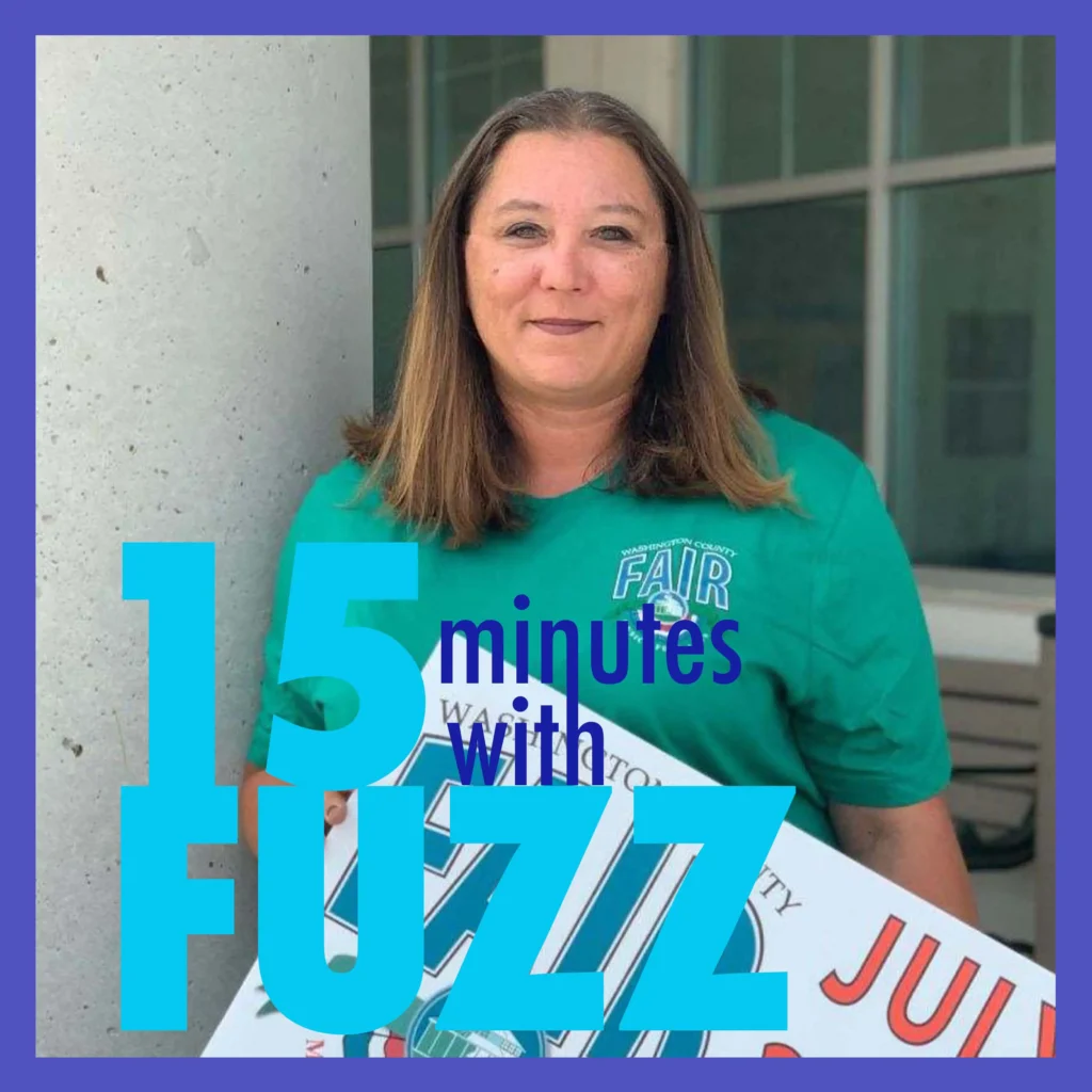 A woman with shoulder-length brown hair stands in front of a building, wearing a green shirt with the text “Washington County Fair” on it. She is holding a sign that appears to promote the fair, which includes the date “July” and part of a logo. She is smiling slightly and looking directly at the camera. In the foreground, the text “15 minutes with Fuzz” is prominently displayed in a bold font.